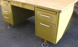 This steel tanker desk boasts serious mid-century modern style. Probably manufactured in the 1950s or 60s. Made to last. Features: 5 drawers, a pencil drawer, pullout writing tablets on each side, and a wood pattern laminate top that overhangs.