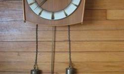 This beautiful mid-century piece offers a modern take on the classic pendulum wall clock. This is a "Perfecta" model from the 1950/60's. Watch case is made of wood, perhaps teak. The swinging pendulum and weights appear to be brass. I believe its calming